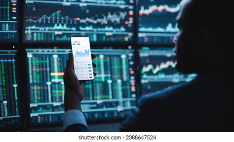 Financial Analyst Using Smartphone with Real-Time Stocks, Commodities and Market Charts. Working on Multi-Monitor Workstation with Stock Charts. Businessman Works in Investment Bank in the Evening. - Powered by Shutterstock