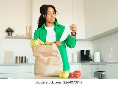 Financial Crisis. Concerned Black Lady Buyer Holding Bill Unpacking Shopper Bag With Products After Grocery Shopping Standing In Modern Kitchen Indoors. Rising Food Prices, Inflation Concept - Powered by Shutterstock