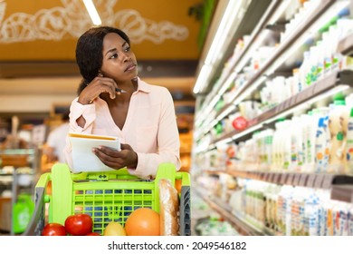 Financial Crisis. Unhappy African American Woman Holding Grocery Shopping List Looking At Shelf Calculating Prices In Modern Supermarket Buying Food Products. Lady Posing Standing With Shop Cart