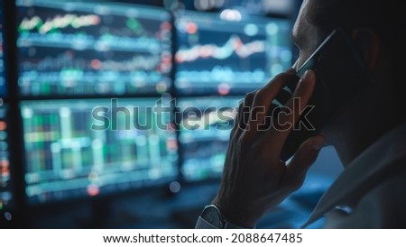 Financial Day Trader Working on a Computer with Multi-Monitor Workstation with Real-Time Stocks Charts and Talking on a Phone with a Client, While in Office Late in the Evening. Close Up Focus.