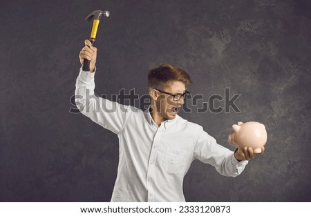 Financial difficulties. Emotional and crazy frustrated young man is going to break piggy bank with hammer. Angry man shouting holding piggy bank and hammer in his hands standing on gray background.