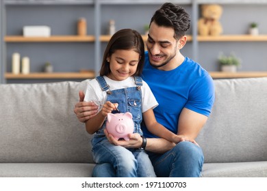 Financial Education For Children Concept. Portrait of smiling little girl putting coin in pink piggy bank, sitting on dad's lap on the couch at home, man teaching his daughter to invest - Shutterstock ID 1971100703