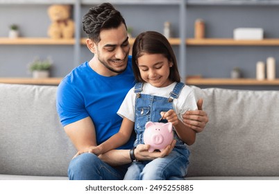 Financial Education For Children Concept. Portrait of smiling little girl putting coin in pink piggy bank, sitting on dad's lap on the couch at home, man teaching his daughter to invest - Powered by Shutterstock