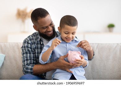 Financial Literacy. Cheerful African Father And Son Putting Money In Piggybank Sitting On Sofa At Home. Daddy Teaching His Child Budget Planning, Keeping Personal Savings Safety