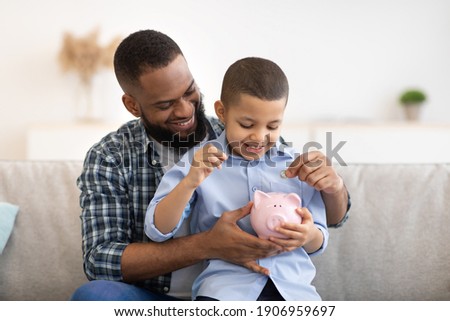 Financial Literacy. Cheerful African Father And Son Putting Money In Piggybank Sitting On Sofa At Home. Daddy Teaching His Child Budget Planning, Keeping Personal Savings Safety