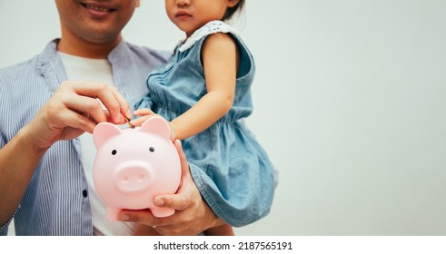 Financial Literacy. Cheerful Asian Father And Daughter Putting Money In Piggybank While Relaxing In Living Room At Home. Money Saving Concept.