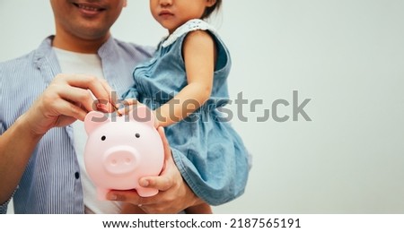 Financial Literacy. Cheerful Asian father and daughter putting money in piggybank while relaxing in living room at Home. Money Saving Concept.