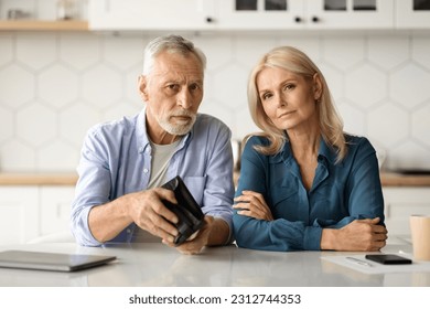 Financial Problems. Upset Senior Couple Showing Empty Wallet While Sitting At Table In Kitchen, Mature Husband And Wife Suffering Retirement Crisis, Having Economy Isuues, Looking At Camera