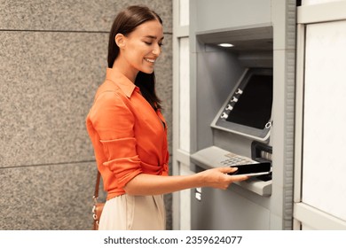 Financial transactions, money withdrawal. Smiling lady putting phone to ATM machine reader, withdrawing cash from bank account in urban area outside. Customer uses smartphone to gain access to ATM