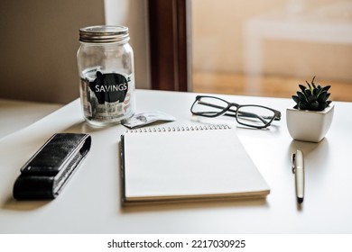 Financial Wellness. Open Empty Notepad Reedy For Planning On Table With Mason Jar Saving Bank, Stationery And Glasses. Budgeting, Investment, Life Insurance, Retirement Plan Concept