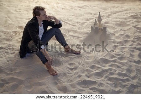Financier and sand castles, the concept of unprofitable investments. Economic crisis and recession. A young man in a business suit is sitting on the sand next to a sand castle