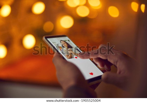 Find love online concept. Man holding mobile phone,\
looking at attractive young woman\'s profile photo on dating app and\
pressing red heart like button. Close-up, blur, romantic bokeh,\
soft focus