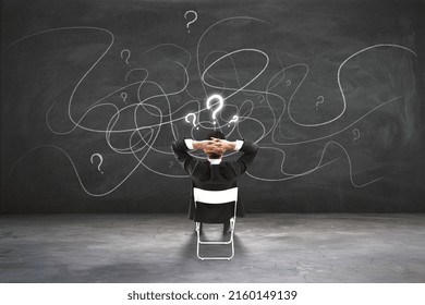 Find solution and making choice concept with back view on businessman sitting on chair and looking at blackboard with question marks and chaotically written lines