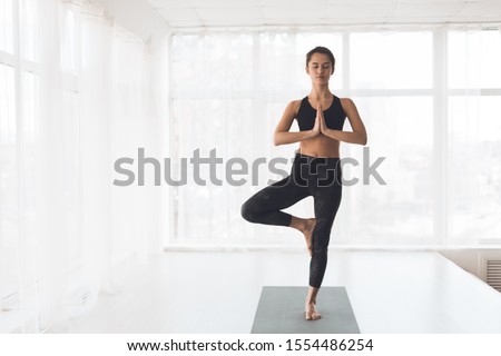 Finding right balance. Woman doing perfect tree pose in light studio against window, free space