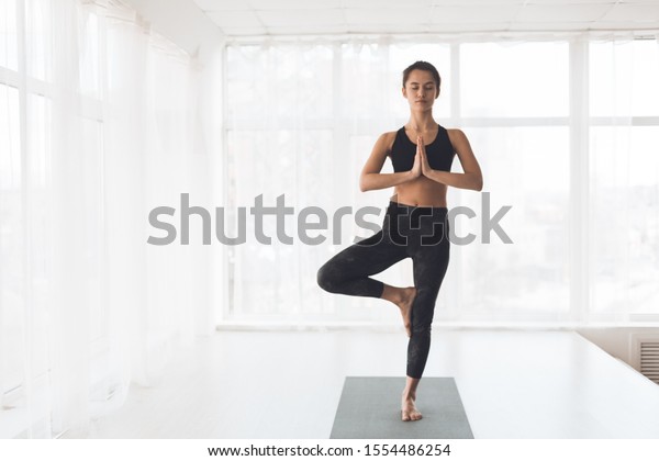 Finding right balance. Woman\
doing perfect tree pose in light studio against window, free\
space