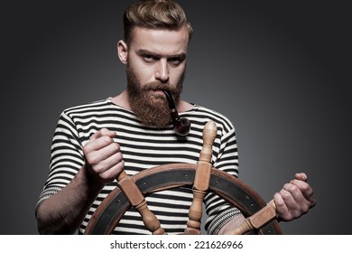 Finding The Right Way. Confident Young Bearded Sailor Steering A Wheel While Standing Against Grey Background 