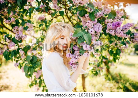 Fine art portrait of young blonde girl in white elegant dress in summer blooming lilac garden. Colorful bushes with flowers on branches. Beautiful cinderella fahion style. Pretty woman at nature