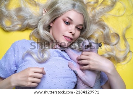 Fine art portrait young blonde woman cosplay elf in blue dress with eyes of different colors lying down on yellow background, holding playful Sphinx kitten on her breasts. Elf has nice curly long hair