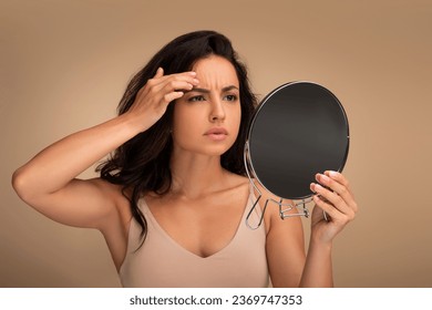 Fine lines and wrinkles, premature skin aging concept. Concerned upset young eastern woman looking in mirror, touching her forehead, checking aging signs on her face, beige background