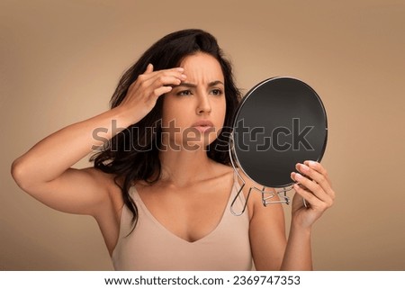 Fine lines and wrinkles, premature skin aging concept. Concerned upset young eastern woman looking in mirror, touching her forehead, checking aging signs on her face, beige background