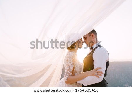 Fine-art destination wedding photo in Montenegro, Mount Lovchen. The wedding couple hugs on top of the mountain at sunset, the groom hugs the bride by the waist, the veil waved into the air.