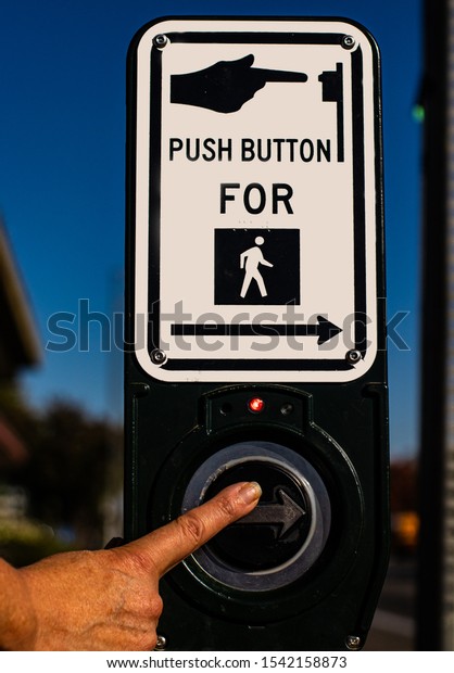 Finger of older person pushing walk signal\
button at a crosswalk