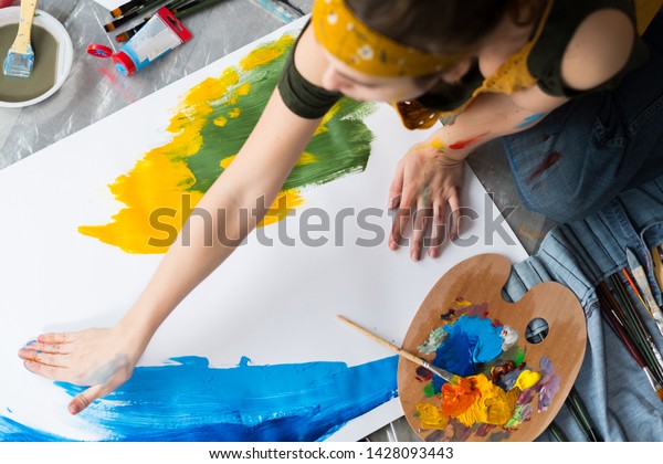 Finger\
painting hobby. Top view of female artist sitting on floor, using\
blue and yellow colors to create abstract\
artwork.