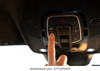 Finger pressing interior light button on overhead console in modern car, close-up of roof control panel with illuminated cabin lighting control.