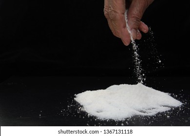 In The Fingers Of An Elderly Woman A Pinch Of Salt, Which She Pours On A Hill Of Salt. Image On Black Background. Concept - Salt-free Diet