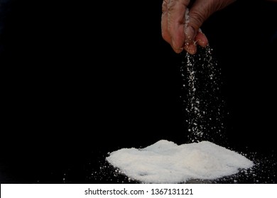 In The Fingers Of An Elderly Woman A Pinch Of Salt, Which She Pours On A Hill Of Salt. Image On Black Background. Concept - Salt-free Diet