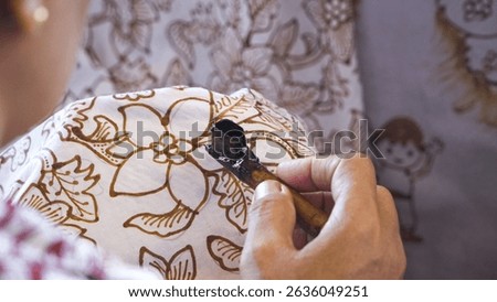 The fingers of a female artist with a batik canting, with a clear close-up shot painting a flower pattern on a unique Balinese batik cloth, she looks very careful and patient.