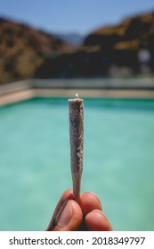 Fingers holding a handmade Marijuana joint (cannabis cigarette) in front of a water green pool between the hills	