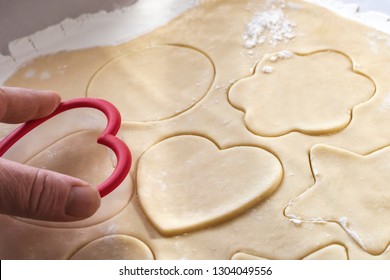 Fingertips Pressing A Heart Shaped Cookie Cutter Into A Sheet Of Sugar Cookie Dough.  Cut Cookie Shapes Include:  Heart, Flower, Star, And Circle.  Close Up Side View.