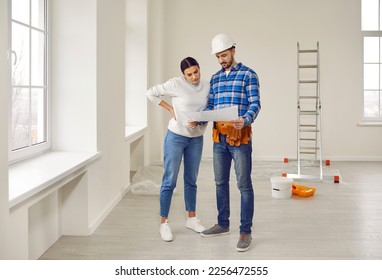 Finishing painting decorating work foreman builder supervisor in hardhat showing young woman homeowner renovation plan standing in empty house or apartment with ladder and plaster bucket in background - Shutterstock ID 2256472555