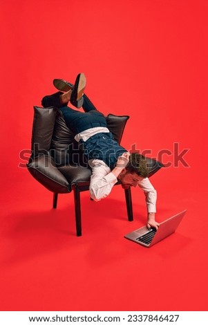 Finishing tasks. Businessman lying on armchair in weird pose, working on laptop against red studio background. Concept of business, working routine, deadlines, freelance, office, ad