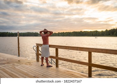 Finnish sauna. Sunset on the lake. Wooden bathhouse or sauna with descent into the water. A man in a special hat for a bath is wrapped in a towel. He looks at the sunset and raises his hands up.