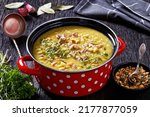 Finnish split pea soup Hernekeitto with ham on red pot with hot mustard and pumpkin seeds on dark wood table, horizontal view from above, close-up