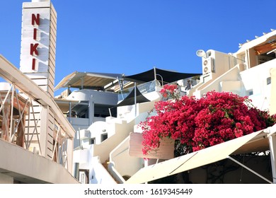 Fira, Santorini in Greece - June 15, 2017: A scene of villas, bars and restaurants in the town of Fira with a large bougainvillea plant and two people on a terrace in the background.