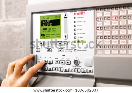 Fire alarm control panel is activated and in alert mode. Display message: Alarm active hall smoke. Red flickering lights and peeping. A hand is using the silence button. Selective focus.