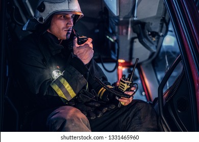 The Fire Brigade Arrived At The Night-time. Serious Fireman Sitting In The Fire Truck And Talking On The Radio