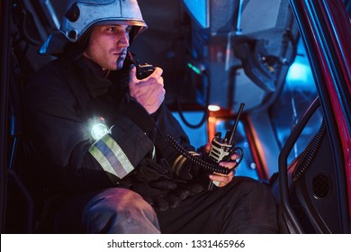 The Fire Brigade Arrived At The Night-time. Serious Fireman Sitting In The Fire Truck And Talking On The Radio