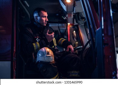 The Fire Brigade Arrived At The Night-time. Serious Fireman Sitting In The Fire Truck And Talking On The Radio