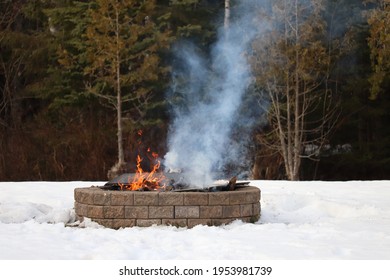 Fire Burning In An Outdoor Firepit In The Winter. Smoke Rising From A Bonfire In A Brick Fire Pit Surrounded By Snow 