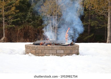 Fire Burning In An Outdoor Firepit In The Winter. Smoke Rising From A Bonfire In A Brick Fire Pit Surrounded By Snow 