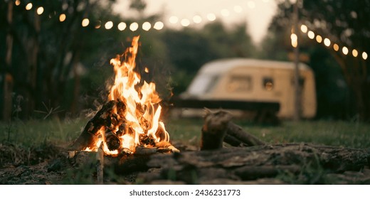 A fire is burning in a fire pit in a grassy area. The fire is surrounded by logs and there are people standing around it. Scene is warm and inviting