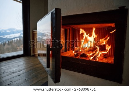 Fire burns in modern fireplace inside cozy home. Close up of glowing logs and flames, glass door open, wooden floor and mountain view through window, warmth and comfort concept