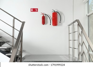 Fire Extinguishers And Emergency Exit Sign On White Wall Near Staircase Indoors