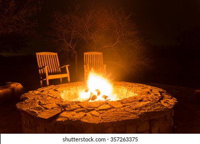 Fire flame in a fire pit and two empty wood chairs next to it with dark forest at the back