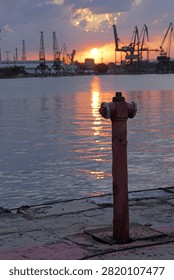 Fire hydrant standing on a concrete pier overlooking an industrial harbor with cargo cranes silhouetted against a dramatic sunset sky, reflecting vibrant orange and purple colors on the calm water