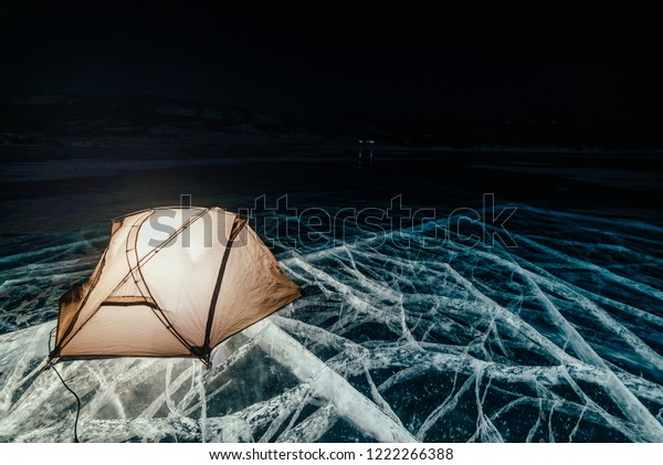 Fire on ice at night. Campground on ice. Tent stands\
next to bonfire. Lake Baikal. Nearby there is car. Shelter tent and\
ice are illuminated from the inside. Beautiful bonfire on real\
cracked ice